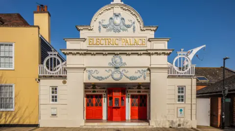 Edwardian cream-coloured building with red doors and Electric Palace in bas relief gold lettering above the door. The building is decorated with plasterwork of wreaths and vines, painted grey. 
