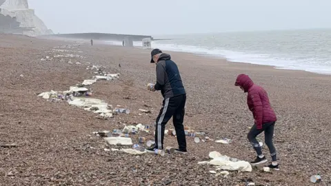 Dan Jessup Two people wearing jackets stood on a line of debris deposited along a beach. The debris includes white foam and small yellow tins. A damaged shipping container and a white cliff can be seen in the background, further down the beach.