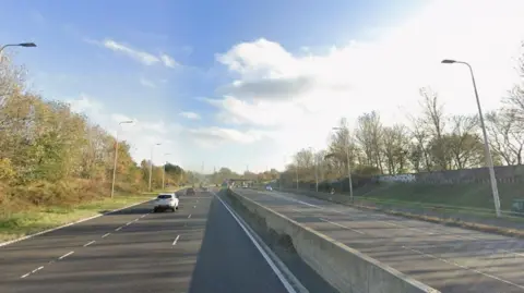 The A19 between North Shields and Wallsend looking south as it heads towards the Tyne Tunnel. It is a dual carriageway with three lanes on either side of a concrete central barrier. There are street lights on each side and trees set back from the road. A wall with graffiti sits on top of a low grass embankment on the northbound side. There is light traffic in both directions and the sky is blue with sun creeping through clouds.