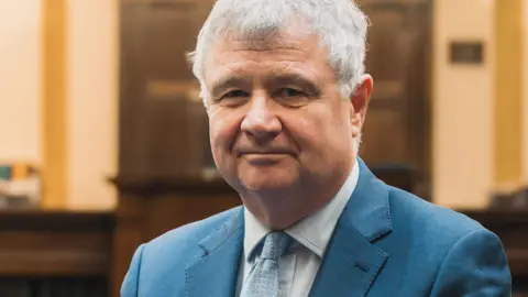 IOM GOVERNMENT Chris Thomas, who has short grey hair and is wearing a blue suit and tie. He is standing in the Tynwald Chamber and there is wooden panelling behind him.
