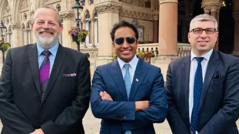 James Petter with short grey hair and a beard, Zia Yusuf with short dark hair and dark glasses, and Mark Arnull with short grey hair and glasses all smiling at the camera and wearing suits. The stone pillars of Northampton Guildhall are behind them.