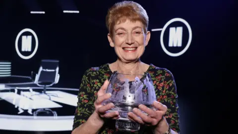 Diane Howe with the Mastermind glass bowl trophy. She has short ginger hair and is wearing a black, green and red flowery dress.