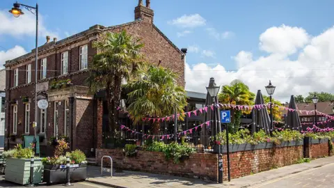 1936 Pubco Exterior view of the Old School House. It is a large red-brick building with a garden full of greenery and bunting. The photograph was taken on a sunny day.