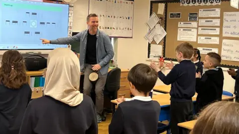 A male teacher with brown hair and a black t-shirt is pointing to a whiteboard with young children in the room clapping. 