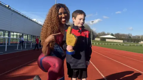 Jake and Montell Douglas are on a running track. Montell is pointing at Jake, who is holding an award.
