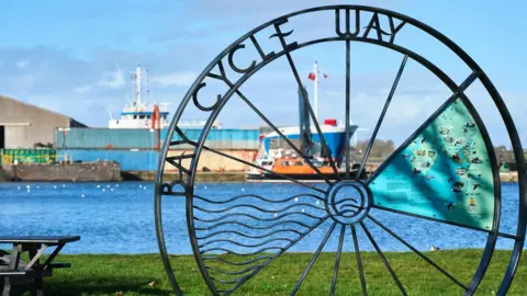Wildey Media Bay Cycle Way marker at Glasson Dock. It is in the shape of a wheel, with a inside and Bay Cycle Way written across the top left. There are benches and some ships in the background.