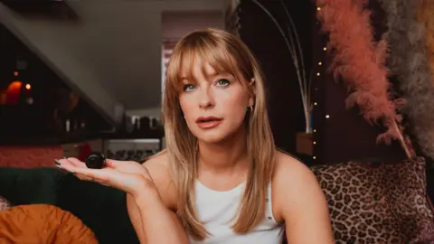 A blonde woman with a fringe looking directly at the camera whilst holding a small black spy camera in her right hand. She's also wearing a white top. 