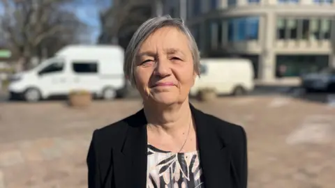 A woman wearing a black blazer and floral top smiles into the camera. She is standing outside on a sunny day. Buildings and parked vans are in the blurred background behind her.