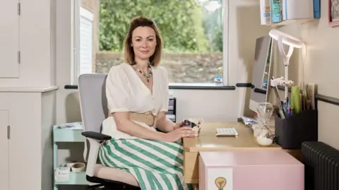 Handout Gee is wearing a white blouse and white and green striped skirt. She is sat at a work table in front of a computer in what looks like an office. There is a window behind her. She has short hair and is smiling at the camera.