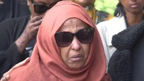 Mr Abdi Mohamed's mother Zahra, who is a Somali woman wearing an orange headscarf and sunglasses. She is crying