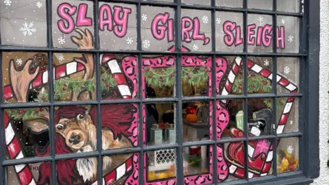 Debbie Carr A shop window that has been painted. It depicts a snowy scene with a reindeer, who has a flowing red wig on. The reindeer is standing inside a red and white candy cane patterned picture frame. There is a pink cheetah print picture frame in the middle, and other candy cane picture frame on the right. Inside this one is a sleigh full of presents. Above the scene in pink lettering, it reads "slay girl sleigh"