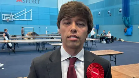 Matthew Webb/BBC Cameron Holloway standing in a sports hall with rows of empty tables behind him. He has mid brown hair in a fringe over his forehead and is wearing a grey suit over a white shirt and maroon tie, with a red Labour rosette on its left lapel.