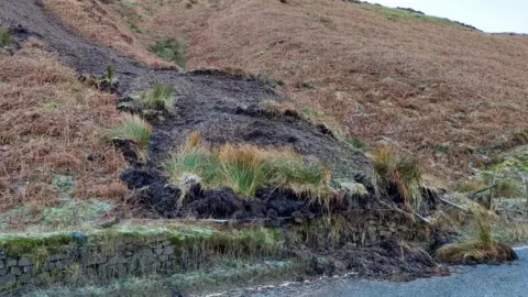 North Yorkshire Council The landslip on the A59 at Kex Gill - a road with a dry stone wall and earth shown falling over the top onto the road 