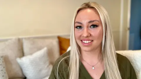 Nikki Fox/BBC Hollie Thornill looks at the camera. She is sitting on a chair in front of the sofa in her living room. She has bleached blonde hair, parted in the middle and is smiling. She has a khaki green blouse on with a crystal necklace. Her sofa is cream, with cream cushions, the wall is beige.