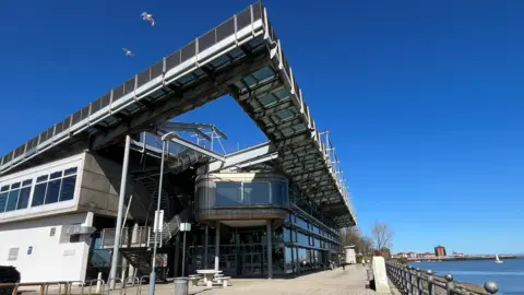 The National Glass Centre is a tall building made of metal panels and glass, overlooking the River Wear. There is seating space and a cafe in front of it, with a dog walker in the distance.
