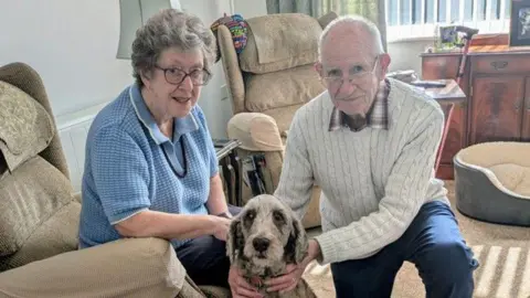 Maureen and Richard Elmer with Gracie the poodle-cross. On the left is Maureen who is sitting on a chair in a living room. She is wearing a blue knitted top with a necklace and is wearing a pair of glasses. She is looking directly at the camera and smiling. On the right is Richard who is kneeling down with his arms around Gracie. He is wearing a red and white checked shirt with a cream jumper over the top and a pair of glasses. He is looking directly at the camera and smiling. In the middle of the couple sat on the floor is Gracie, a poodle-cross with light brown fur.