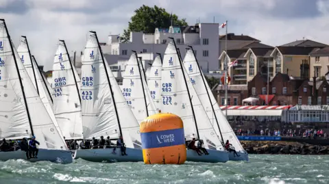 North Sails Cowes Week A group of yachts, all with white sails, lined up with Cowes in the background and a yellow marker bouy in the sea in the foreground