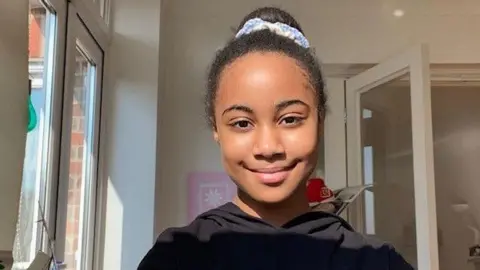 Daisy smiles at the camera inside her house. She wears a black hooded top and has her hair tied back into a bun on her head, with a blue scrunchie.