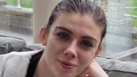 Holly Collinson is seen with her hair scraped back, an eyebrow earring and she smiles toward the camera. 