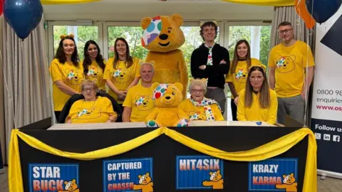 The Orders of St John Care Trust A team photo of care home residents and staff all wearing yellow, with Pudsey and Darragh Ennis