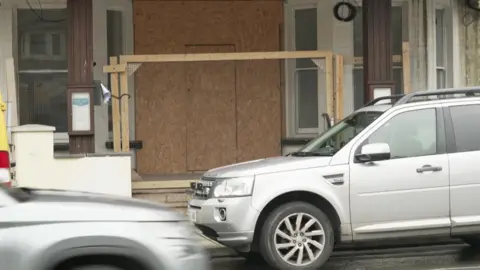 The pictures is a street‑level shot of the front of a building that has been boarded up. The entire doorway area is covered with sheets of brown wooden board, and there is a simple wooden frame built in front of it, with horizontal and vertical beams holding everything in place. 