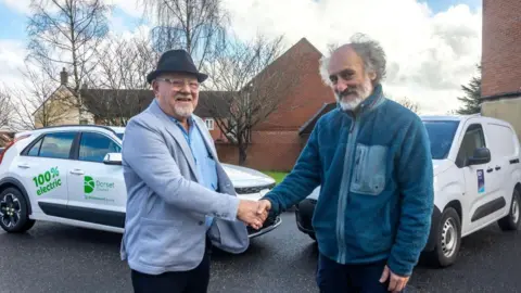 Dorset Council Councillors Jon Andrews and Andy Hadley shake hands as they pose for the camera in front of Dorset Council and BCP-branded electric vehicles. Andrews is wearing a light grey blazer and a black trilby hat and has a grey goatee beard and glasses. Hadley is wearing a royal blue fleece and has grey hair and a full beard.