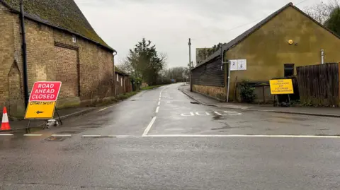 JOHN DEVINE/BBC The B1049, a 'T' junction with no traffic visible at all, but temporary signs each side of the roadway saying 'Road ahead closed' and 'Diversion' with a black arrow pointing left, a second yellow sign opposite says 'B1049 closed ahead for Cottenham, follow diversion. Some buildings can be seen either side of the road.