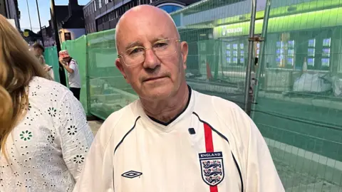 Phil Harrison/BBC A bald man with glasses wearing a white England football shirt.