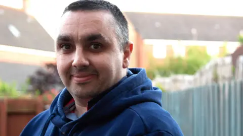 East Anglian Air Ambulance Jonathan Moran is standing in a garden which has fences around it. He is wearing a navy hooded jumper and has short black hair. There are rows of houses in the background.