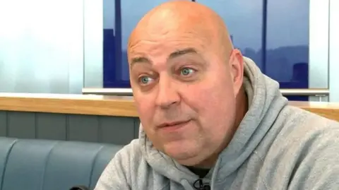 A still of Mark Garland with a bald head and blue eyes wearing a grey hoodie talking to someone off camera inside a room