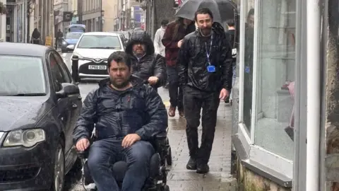 Access Cornwall A man uses a wheelchair on the narrow pavements in Bodmin , he is soaked because it's raining hard.