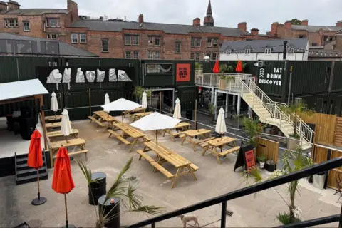 An open yard with shipping containers, stairs and seats with umbrellas.