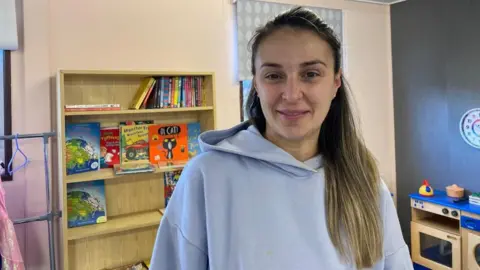 Ilia Pascu smiles at the camera, standing in a children's playroom. She has long, dark blonde hair and wears a light grey hoodie. In the background, a wooden shelf can be seen with children's books, and a wooden kitchen play set.