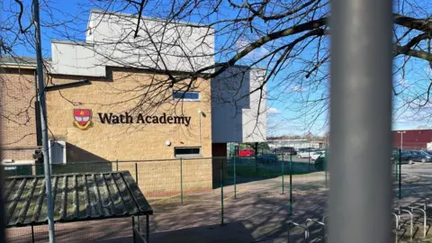 A photo of a sandstone building with "Wath Academy" on the side. There is a car park to the right of the building and what looks like a bike shelter to the foreground.