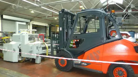 HSE A large orange forklift holding a grey machine in place. Safety tape is around both of them. 