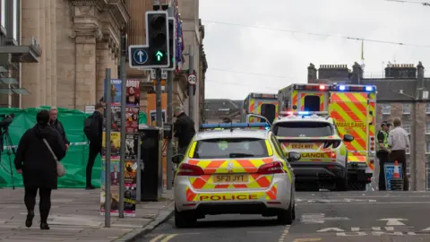 two police cars and an ambulance outside a budling, a green cordon is visible to the left.