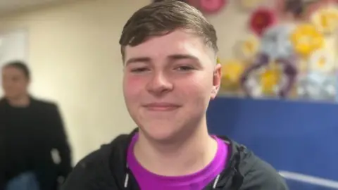  Riley is wearing a black Nike jacket and bright purple top. He is smiling at the camera and has brown hair.