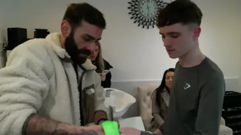 Matthew Wood, in a white woollen fleece, places a green sensory wristband around the left arm of 13-year-old Harry, who is wearing a green long-sleeved top