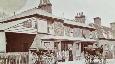 The Queen Victoria An old sepia coloured photo of the pub from outside. There are two horse drawn carriages but no horses. Four people can be seen stood outside in Victorian dress looking at a carriage.