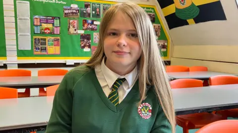 A girl with blonde hair looking at the camera. She has a green jumper on, over a white shirt and a green and yellow striped tie.