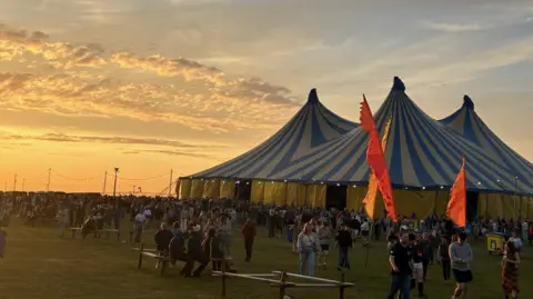 Rachel Butchert Large striped festival tent at sunset with crowds gathering.