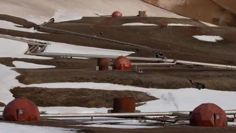 Steel pipes connect the red pods of a geothermal power station in the Icelandic landscape.