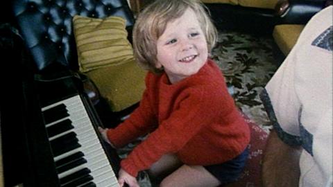 Toddler Vincent Billington, dressed in a red jumper, sitting at a piano and smiling.