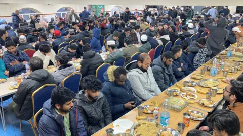 Rows of people, most of them men, are sitting in front of tables which are loaded with food. The setting is a mosque and some of the men are wearing the short brimless hat worn by some Muslims.  