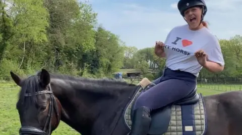 A woman, Tasha Lee Hopkins, sits on a horse pointing to her T-shirt, which reads: I love my horse. She is wearing dark blue jodhpurs, black riding boots and a black riding helmet. Her hair is tied up. She is sat on a dark brown horse that is stood in a field.