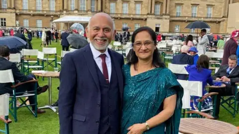 George Miah This image shows two people standing outdoors at a formal event on a grass lawn, surrounded by rows of folding chairs and small tables. One man is wearing a dark suit with a tie, while a woman is dressed in a teal, draped formal outfit. In the background, there is a large historic building with stone architecture and a domed roof, and several other attendees are visible, some holding umbrellas, suggesting unsettled weather. The setting appears ceremonial or celebratory, with organised seating and people socialising.