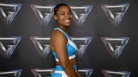 Emily Bell in a blue Gladiators kit, stands in front of the Gladiators branded wall