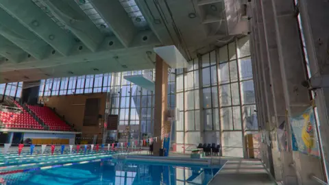Aaron Law An indoor swimming pool with large glass windows on one side and rows of red seats on the other
