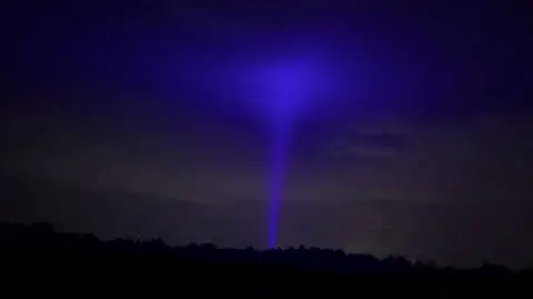 A bright blue hue in the dark sky, resembling a tornado-type shape, with a narrow stem and wider top. Dark trees can be seen in the foreground.