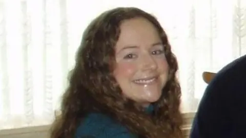 A young women with brown wavy hair is looking at the camera and smiling.  She is wearing a blue jumper